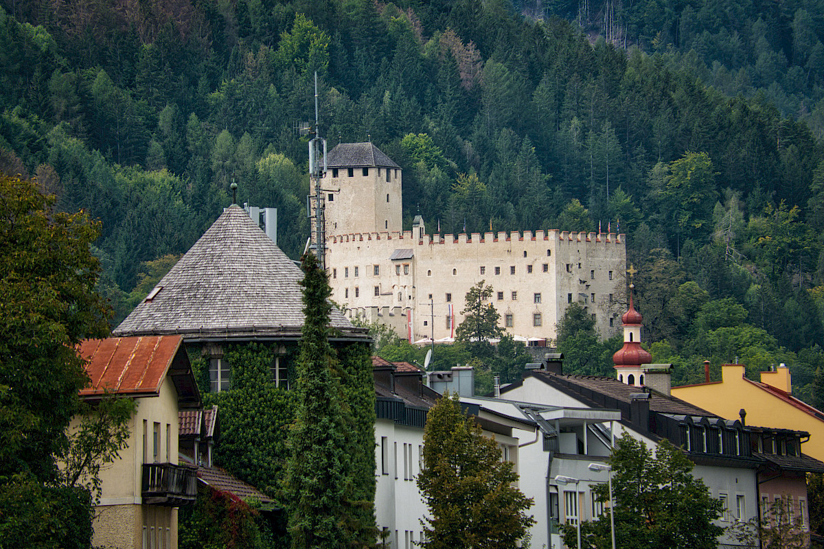 Eine mittelalterliche Burg vor bewaldeten Bergen und Häusern einer Kleinstadt.